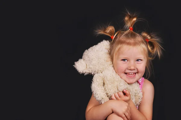 Chica feliz con colas en la cabeza sosteniendo un oso de peluche y sonriendo sobre un fondo negro — Foto de Stock