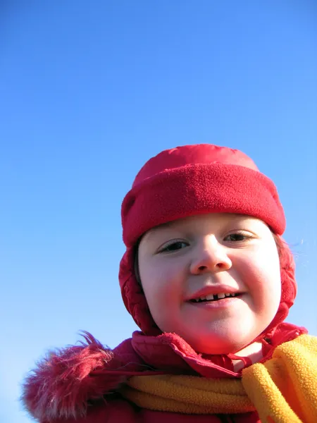 Klein meisje in een rode muts met oorkleppen op blauwe hemelachtergrond lachen — Stockfoto