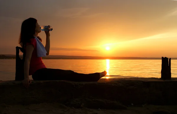 Schöne junge sportliche Frau, die vor einem Sonnenuntergang sitzt und trinkt. — Stockfoto