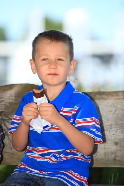 Chłopiec siedzi na ławce w parku, jedzenie czekolady — Zdjęcie stockowe