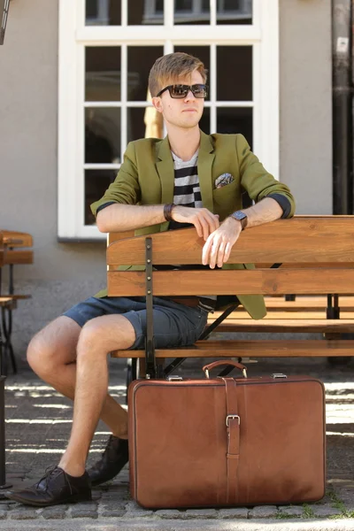 Young handsome man with suitcase waits on bench — Stock Photo, Image
