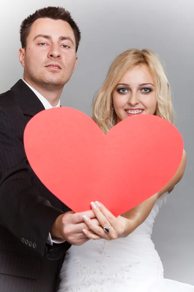 Portret van gelukkige bruid en bruidegom met rood hart op grijs — Stockfoto