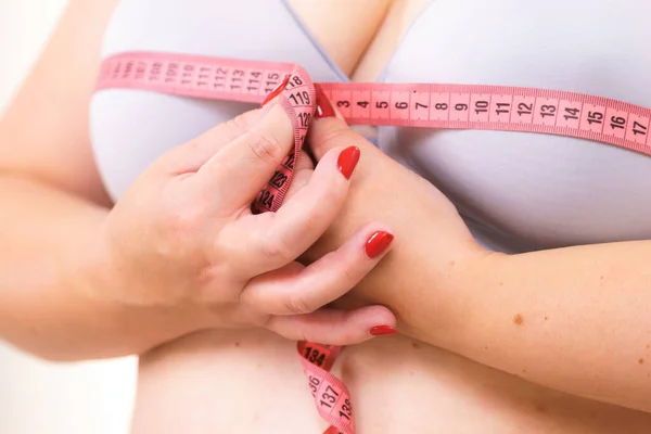 Closeup of female breasts. Plus size fat woman wearing bra using tape measure to check the measurements of her big chest. Bosom, brafitting and underwear concept.