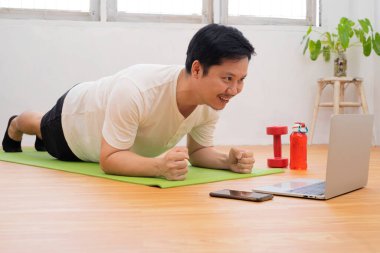 Evde antrenman. Sportif adam dizüstü bilgisayarda ders izlerken yoga tahtası yapıyor oturma odasında egzersiz yapıyor, boş yer var.
