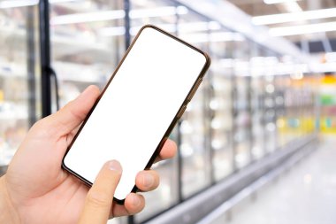 Süpermarkette beyaz ekranda akıllı telefon kullanan bir adam. Fiyat karşılığı süpermarketin bulanık arkaplan ve yeşil filtresi; çevrimiçi alışveriş kavramı