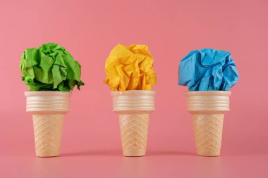 Green, yellow, blue paper cubes placed inside an ice cream cone on a pink background.