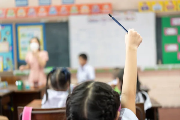 Selective focus on hand. Children or Schoolkids or students raising hands up with Asian teacher wearing protective face mask in classroom at school while covid-19 pandemic. New Normal Life concept.