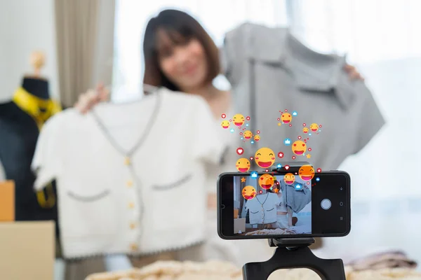 Young Asia lady fashion designer using mobile phone receiving purchase order and showing clothes recording video live streaming online at shop. Small business owner, online market delivery concept.