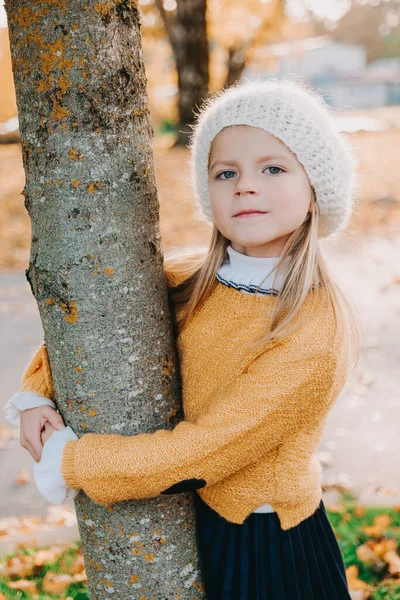 Adorable girl in yellow blouse and white beret hugs the tree