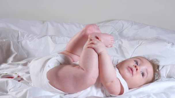 Sevimli 7 aylık bebek yatakta uzanıyor ve bacaklarıyla oynuyor. — Stok video