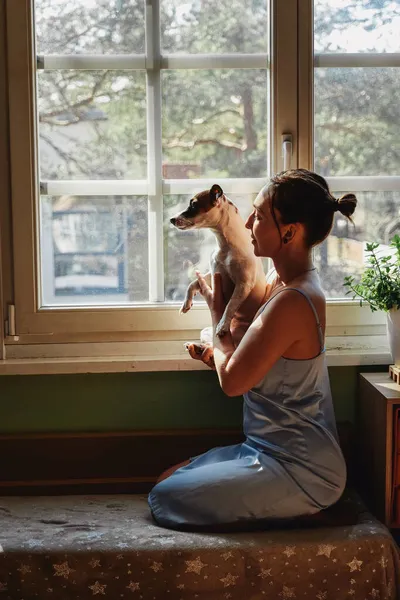 Krásná brunetka žena hladí svého psa u okna — Stock fotografie