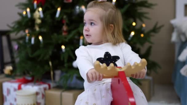 Küçük mutlu kız çocuğunun portresi ahşap geyik üzerinde, Noel ağacı arka planda. — Stok video