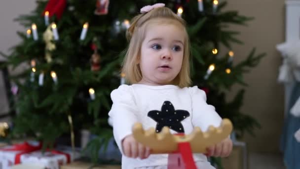 Portré kis boldog gyermek lány a fa rocking-szarvas, karácsonyfa a háttérben — Stock videók