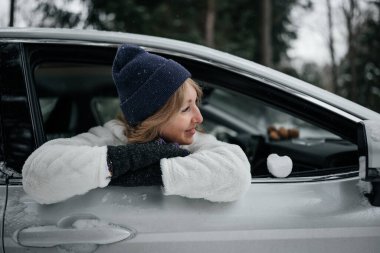Happy woman in stylish winter clothes looking out the car 