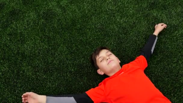 Glad pojke i fotbollsuniform liggande på gräset på fotbollsplanen — Stockvideo