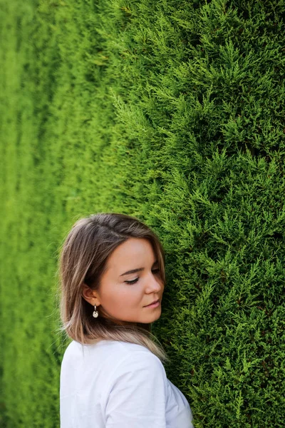 Yeşil çitin yanında duran beyaz gömlekli, doğal makyajlı genç bir kadın. — Stok fotoğraf