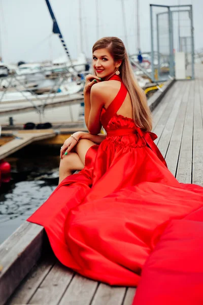 Jeune femme magnifique en robe rouge longue assise sur la jetée — Photo