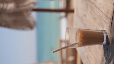 Ice coffee latte on the beach. Summer iced coffee latte in tall glass with straw by the sea in beach bar. Vertical video