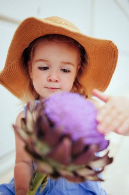 Portrait of cute little 2 years old girl in straw hat touching big artichoke flower. Happy childhood, beautiful kids