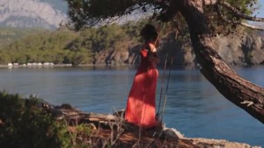 Genç Dinlenen Kadın 'ın arkası turuncu uzun elbisesiyle çam ağacında salıncakta sallanıyor ve güzel deniz manzaralı. Yaz rahatla, sakin güzel tatil köyü. 