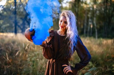 Ormanda, üzerinde mavi duman olan oyulmuş balkabağı taşıyan kahverengi elbiseli güzel genç bir kadının portresi. Cadılar Bayramı partisi, güzel kadın.