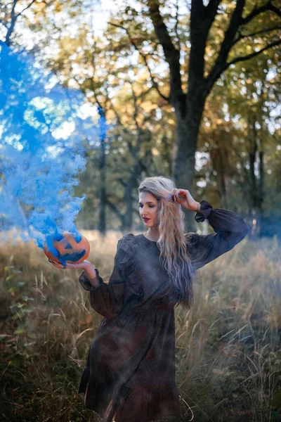 茶色のドレスを保持して美しい若い女性の肖像画は 森の中で そこから出てくる青い煙とカボチャの彫刻 ハロウィンパーティー美しい女性 — ストック写真
