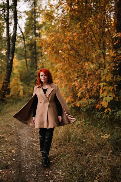 Young beautiful woman with dyed red hair wearing beige cat and leather knee high boots walking happily in autumn forest and looking at camera. Pretty woman revealing individuality.