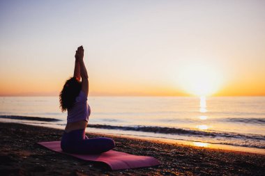 Backside view of calm woman yoga practicing by the sea sunrise time. Finding inner peace spiritual healing lifestyle.