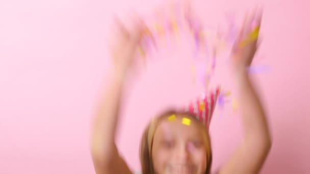 Дівчина-підліток у святковій шапці викидає конфетті на кольоровому фоні крупним планом — стокове відео