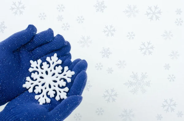 Close-up op handen dragen blauwe handschoenen houden een grote sneeuwvlok. wanten en sneeuwvlok op een witte sneeuw achtergrond. — Stockfoto