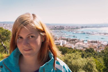 Sarı turuncu saçlı genç bir kadın, Palma ve Majorca 'nın olduğu bir gözlem noktasında dikilirken kameraya bakıyor.