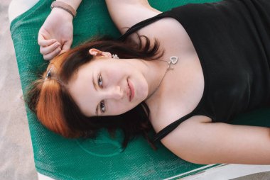 Uzun kırmızı ve siyah saçlı genç bir kadın kumsalda bir iskemlede uzanmış gülümsüyor, kameraya bakıyor, yüksek açılı, yatay manzaralı.