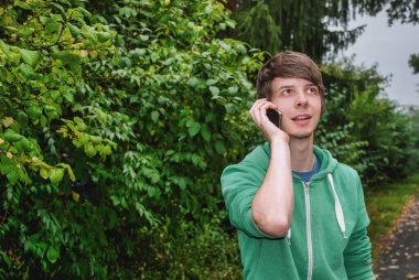 Kahverengi saçlı ve sakallı genç adam ormanda akıllı telefonuyla konuşuyor, yeşil bir kazak giyiyor.