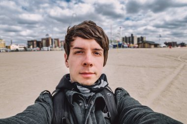 Kahverengi saçlı ve sakallı genç bir adam Coney Island, New York 'un önünde selfie çekiyor. Uçak yorgunluğu, Amerika' nın başkenti