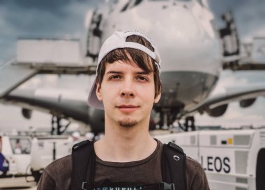 Genç yetişkin bir adam havaalanında büyük bir uçağın önünde dururken kameraya bakıyor.