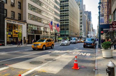 Manhattan, New York, trafiğin yoğun olduğu cadde, sarı taksiler ve diğer özel arabalar, kaldırımda yürüyen insanlar, kahve pastanesi arka planda hazır gıda mağazası, Amerikan bayrakları bir binadan sarkıyor,