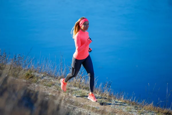 Young sporty woman in orange longsleeve running along blue frosty lake in the morning — Stock Photo, Image
