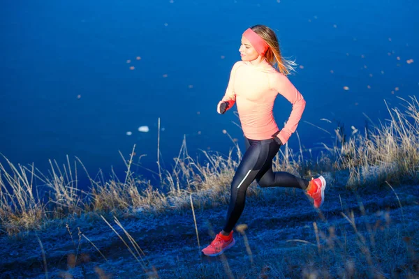 Young sporty woman in orange longsleeve running along blue frosty lake in the morning — Stock Photo, Image