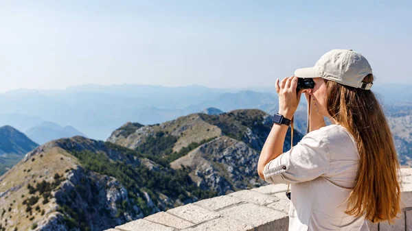 Молодая женщина наблюдает горный ландшафт через бинокль в месте с видом на туристическую тропу — стоковое фото