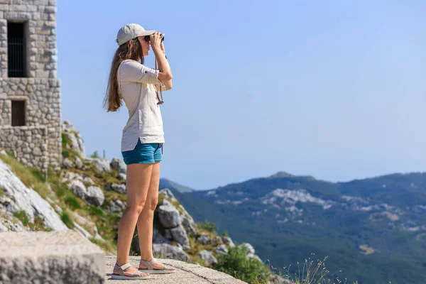 Mujer joven observando el paisaje de montaña a través de prismáticos en el punto de vista en el sendero turístico — Foto de Stock