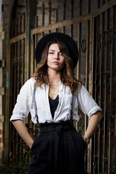 Young woman in white shirt and black hat standing near the gate. Gangster or vintage style girl on dark moody background