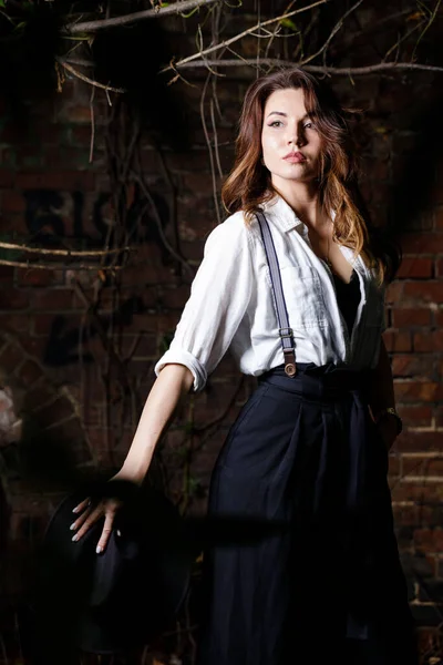 Young woman in white shirt and black hat standing in the darkness. Gangster or vintage style girl on dark moody background