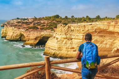 İnce bir adam Portekiz 'de bir kayanın üzerindeki çitin yanında büyük bir sırt çantasıyla duruyor. Arkadan okyanusa bakan bir adam mesafeye bakar.