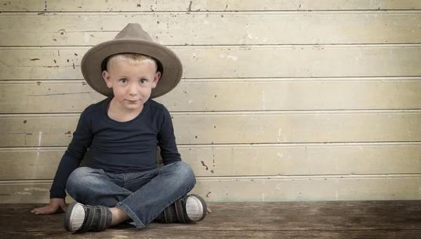 Liten pojke med cowboyhatt som sitter mot en gammal trä dörr — Stockfoto