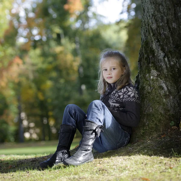 Kleines blondes Mädchen sitzt gegen einen Baum — Stockfoto