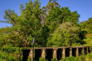 PONTEVEDRA, SPAIN - 1 Mayıs 2021: Maskeli bir adam, bir nehri geçen ilkel granit bir köprüden geçiyor.