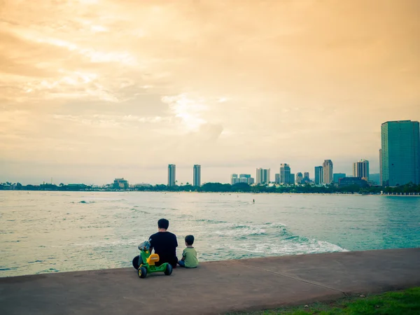 Πατέρας και γιος που κάθονται δίπλα στη θάλασσα με φόντο αστικό τοπίο — Φωτογραφία Αρχείου