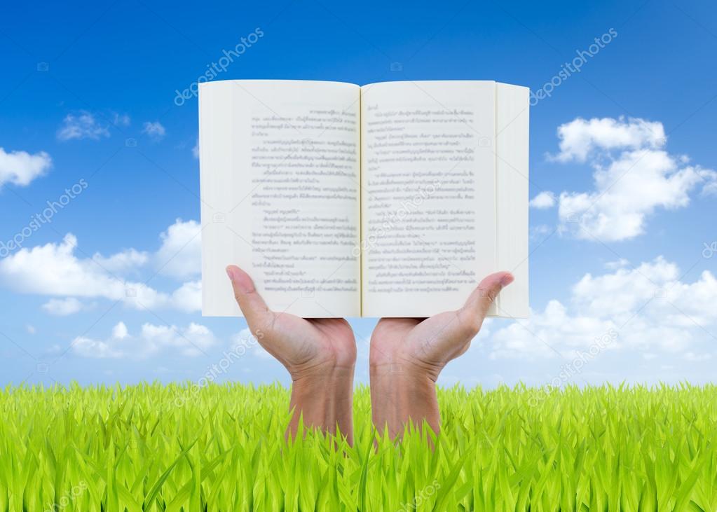 Man hands holding book on green field with blue sky background
