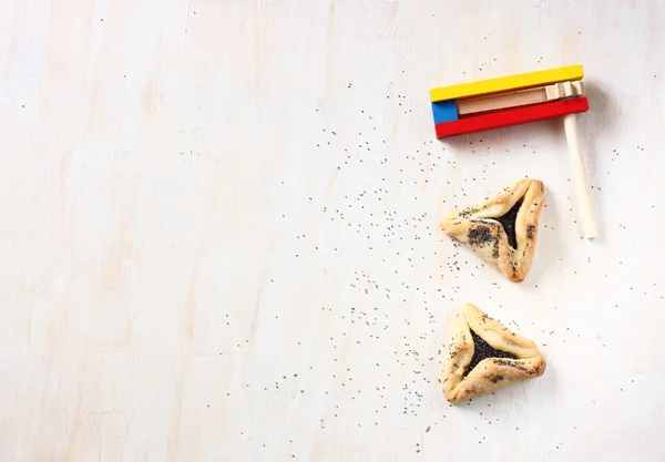 Hamantaschen cookies or hamans ears for Purim celebration and noisemaker over textured wooden board — Stock Photo, Image