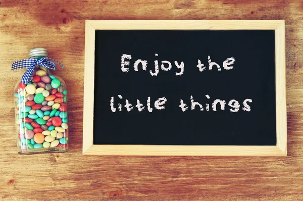 Garrafa cheia de doces e quadro-negro com a frase desfrutar das pequenas coisas . — Fotografia de Stock